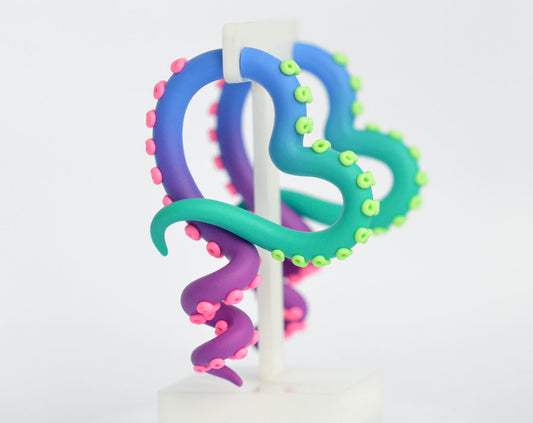 Heart shape octopus tentacle earrings with glow in ultraviolet light suckers. The body of the fake tentacle gauges fades from green to blue to purple but the suckers are neon lime green and neon pink. Under normal lighting, the suckers are light lime green and bubblegum pink.